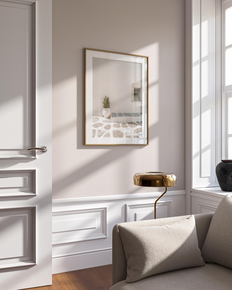 May include: A framed print with a gold-colored frame hangs on a light pink wall. The print depicts a white building with a cactus in a white pot. A gold-colored lamp and a beige sofa are in the foreground.