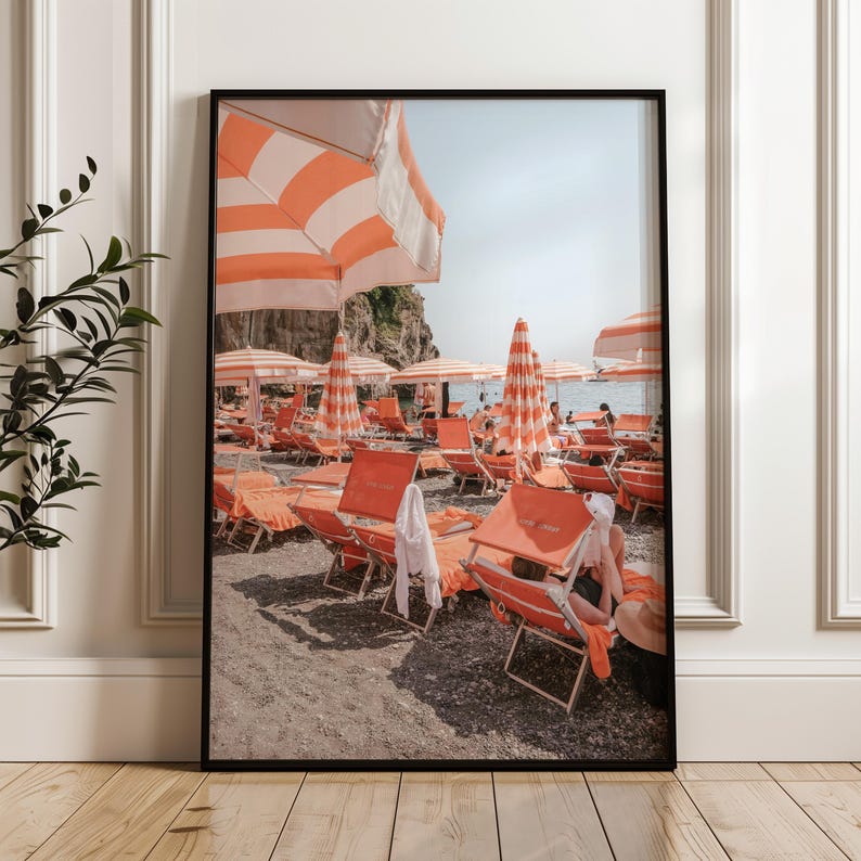 May include: Framed beach scene print featuring orange and white striped umbrellas and lounge chairs. The photograph captures a sunny day at the beach with people relaxing under the umbrellas. The print is in a black frame.