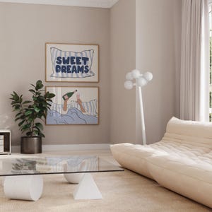 May include: Two framed illustrations on a light beige wall. The top illustration features a blue and white striped pillow with the text "SWEET DREAMS". The bottom illustration shows a person in bed with a glass of champagne and a bottle of champagne. The room has a white sofa and a glass coffee table with a white base.