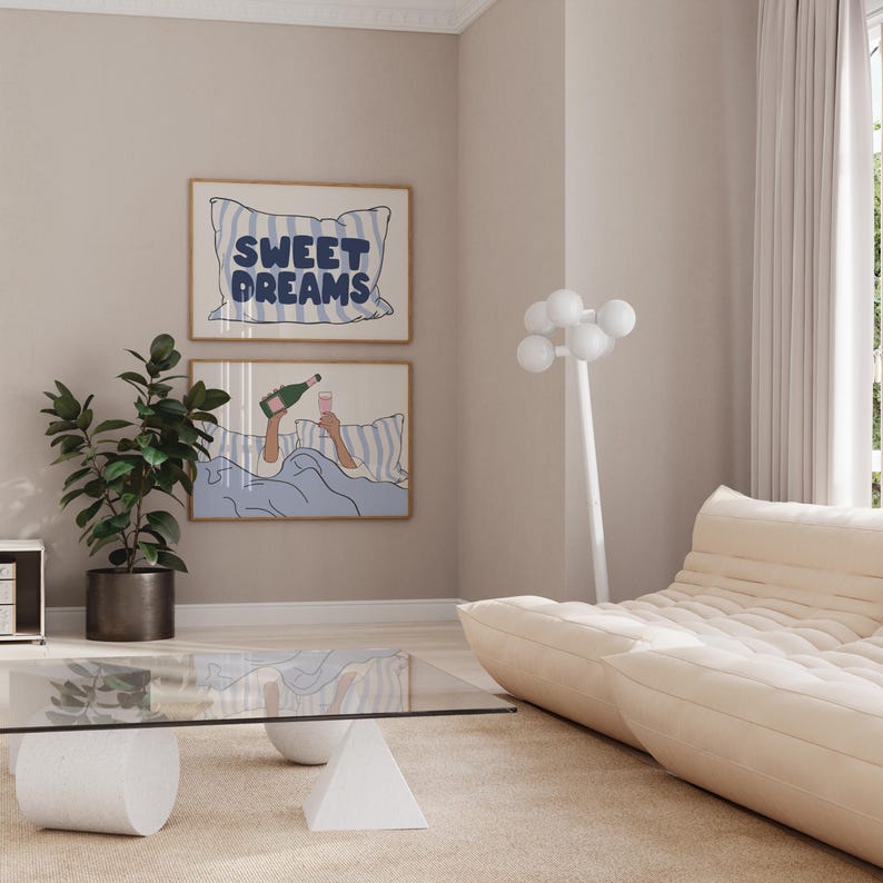 May include: Two framed illustrations on a light beige wall. The top illustration features a blue and white striped pillow with the text "SWEET DREAMS". The bottom illustration shows a person in bed with a glass of champagne and a bottle of champagne. The room has a white sofa and a glass coffee table with a white base.