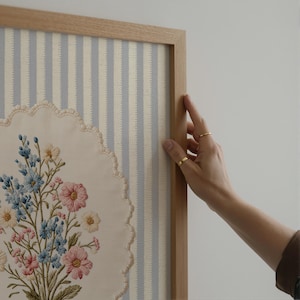 May include: A framed floral embroidery featuring a bouquet of pink, blue, and yellow flowers. The embroidery is set against a striped blue and white background. The frame is made of light-colored wood.