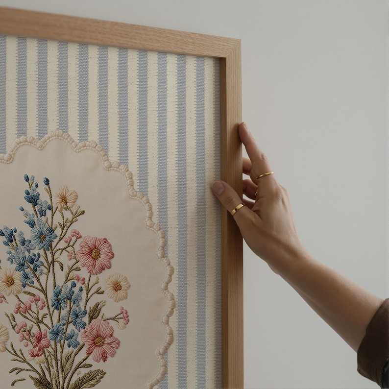 May include: A framed floral embroidery featuring a bouquet of pink, blue, and yellow flowers. The embroidery is set against a striped blue and white background. The frame is made of light-colored wood.