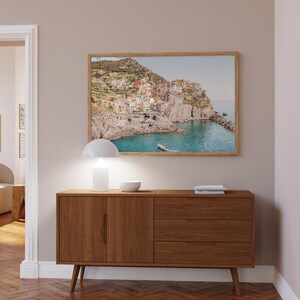 May include: A wooden cabinet with two doors and three drawers, a white lamp, and a framed picture of a coastal town on a wall above the cabinet.