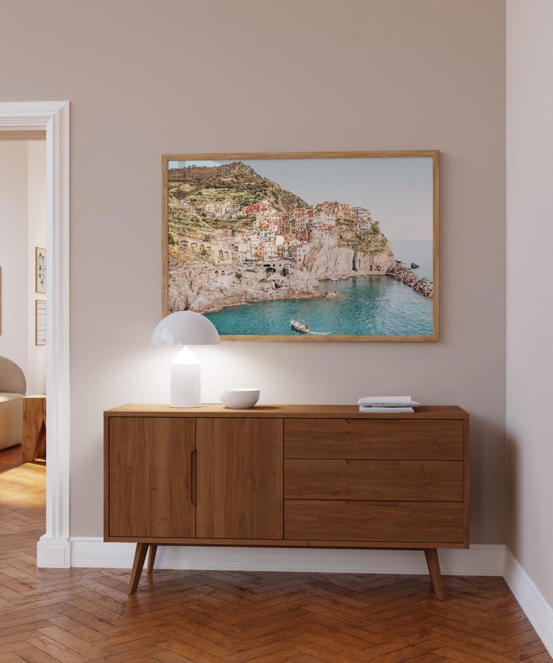May include: A wooden cabinet with two doors and three drawers, a white lamp, and a framed picture of a coastal town on a wall above the cabinet.