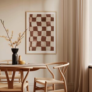 May include: A brown and white checkered abstract painting in a wooden frame hangs on a beige wall above a wooden dining table with two chairs.