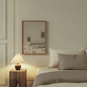 May include: A bedroom scene featuring a framed artwork with a Mediterranean theme. The artwork depicts a white building with a cactus in a vase. A bedside table with a lamp and a bed with pillows and a striped blanket are also visible.