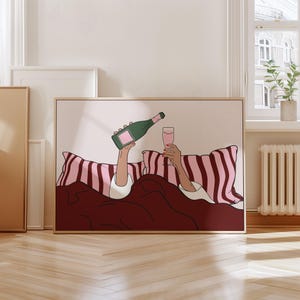 K&ouml;nnte beinhalten: Gerahmter Kunstdruck, der eine Person im Bett zeigt, die Champagner einschenkt. Die Illustration zeigt eine gr&uuml;ne Champagnerflasche, ein rosa Glas und rosa-wei&szlig; gestreifte Kissen. Die Bettw&auml;sche ist dunkelrot.