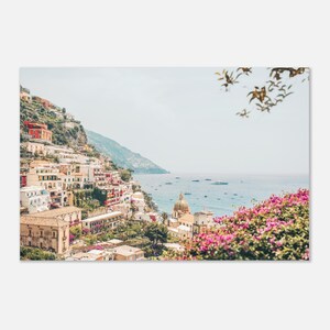 May include: A scenic photograph of a coastal town nestled on a hillside, featuring colorful buildings, a domed church, and the blue sea. Pink flowers frame the foreground, with boats dotting the water under a clear sky.