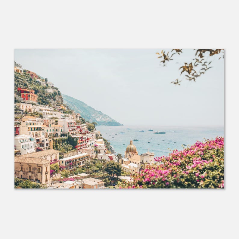 May include: A scenic photograph of a coastal town nestled on a hillside, featuring colorful buildings, a domed church, and the blue sea. Pink flowers frame the foreground, with boats dotting the water under a clear sky.
