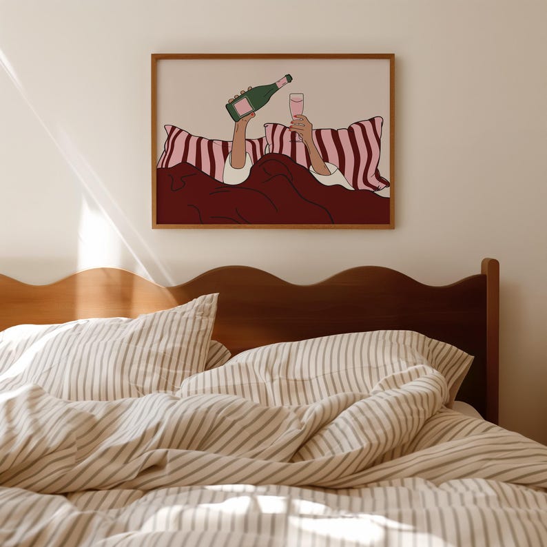 K&ouml;nnte beinhalten: Ein gerahmter Kunstdruck zeigt eine Person im Bett, die eine Champagnerflasche und ein Glas rosa Fl&uuml;ssigkeit h&auml;lt. Das Kunstwerk hat einen braunen Rahmen, einen beigen Hintergrund und eine bordeauxfarbene Decke. Die Kissen haben ein rosa-wei&szlig;es Streifenmuster.