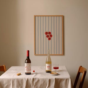 May include: A still life composition with a framed artwork, two wine bottles, a glass of red wine, and a small plate with bread on a table. The artwork features a red cherry design. The table is covered with a white tablecloth.