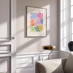 K&ouml;nnte beinhalten: Ein gerahmter Kunstdruck mit einem floralen Design. Das Kunstwerk zeigt abstrakte Blumen in Blau, Rosa, Orange, Gr&uuml;n und Gelb. Der Druck wird in einem Raum mit neutraler Farbgebung, einschlie&szlig;lich eines beigen Sofas und einer goldenen Lampe, ausgestellt.