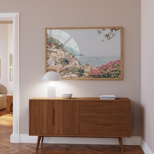 May include: A wooden sideboard with two doors and three drawers, a white lamp, and a bowl on top. A framed print of a coastal town with a blue sky and ocean is hanging on the wall above the sideboard.