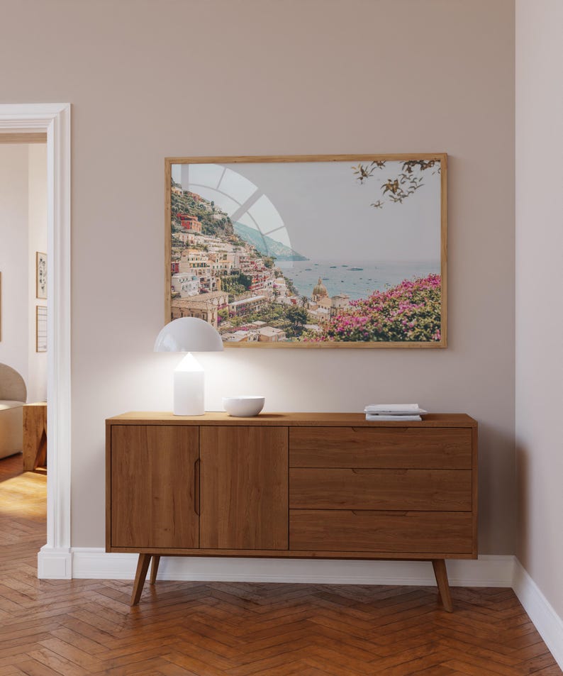 May include: A wooden sideboard with two doors and three drawers, a white lamp, and a bowl on top. A framed print of a coastal town with a blue sky and ocean is hanging on the wall above the sideboard.