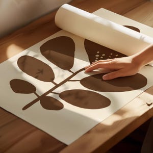 Pu&ograve; includere: Una stampa artistica con un design botanico marrone su sfondo bianco sporco. La stampa &egrave; parzialmente srotolata, rivelando un motivo di foglie e steli. L'immagine &egrave; su una superficie di legno.