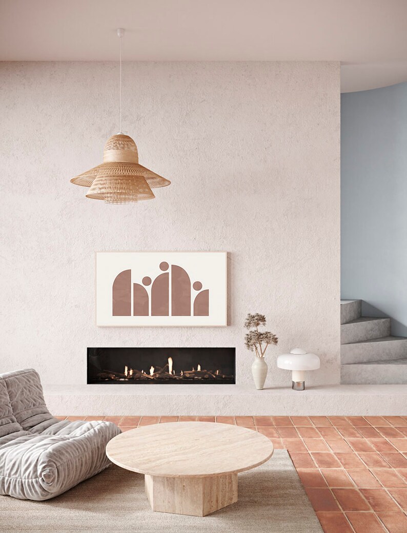 May include: A living room interior with a fireplace, a large, round, light-colored coffee table, a gray couch, a woven pendant light, and a framed print with a geometric design in shades of brown.