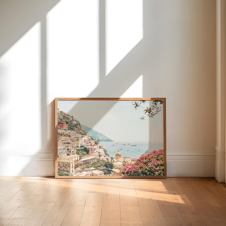 May include: A framed photograph of a coastal town with colorful buildings on a hillside, overlooking a blue ocean with boats. The photo is in a light wood frame and has a white background.