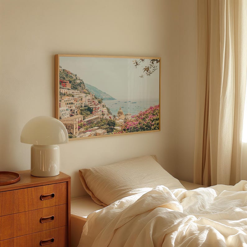 May include: A framed photograph of a coastal town with colorful buildings and a blue ocean in the background. The photo is hanging on a light beige wall above a bed with white linens.