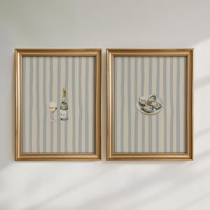 May include: Two framed watercolor prints. One features a wine bottle and glass, the other a plate of oysters. Both have gold frames and a striped background in blue and beige, creating a cohesive set.
