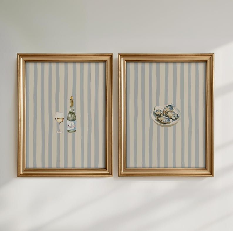 May include: Two framed watercolor prints. One features a wine bottle and glass, the other a plate of oysters. Both have gold frames and a striped background in blue and beige, creating a cohesive set.