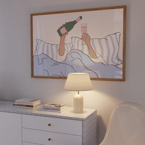 May include: A framed illustration of a person in bed with a glass of champagne and a bottle of champagne. The person is wearing a white shirt and is lying on a blue and white striped bedspread. The illustration is in a light brown frame.