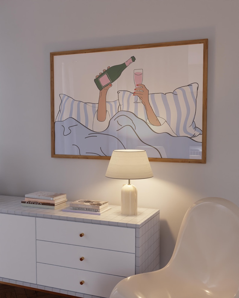 May include: A framed illustration of a person in bed with a glass of champagne and a bottle of champagne. The person is wearing a white shirt and is lying on a blue and white striped bedspread. The illustration is in a light brown frame.