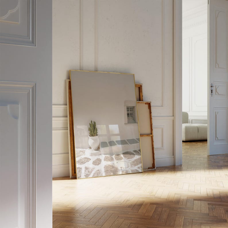 May include: A large rectangular mirror with a gold frame leans against a white wall. The mirror reflects a stone patio with a cactus in a white pot. The scene is set in a room with a wooden floor and white doors.