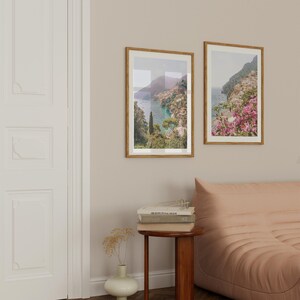 May include: Two framed landscape photographs of a coastal town with a bay and mountains in the background. The photographs are hung on a light beige wall above a pink sofa.