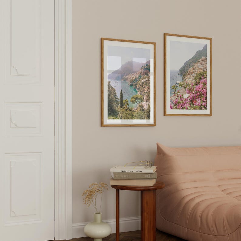 May include: Two framed landscape photographs of a coastal town with a bay and mountains in the background. The photographs are hung on a light beige wall above a pink sofa.