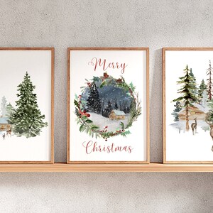 May include: Set of three framed watercolor prints featuring winter scenes. Each print depicts a cabin in a snowy forest with evergreen trees and deer. One print has a wreath with the words "Merry Christmas". The frames are a light brown color.