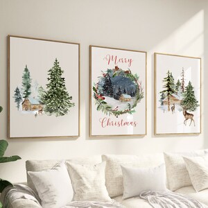 May include: Set of three framed watercolor prints featuring winter scenes. Each print has a gold frame and depicts a cabin, trees, and deer. One print has the text "Merry Christmas" in red script. The artwork evokes a cozy, festive atmosphere.