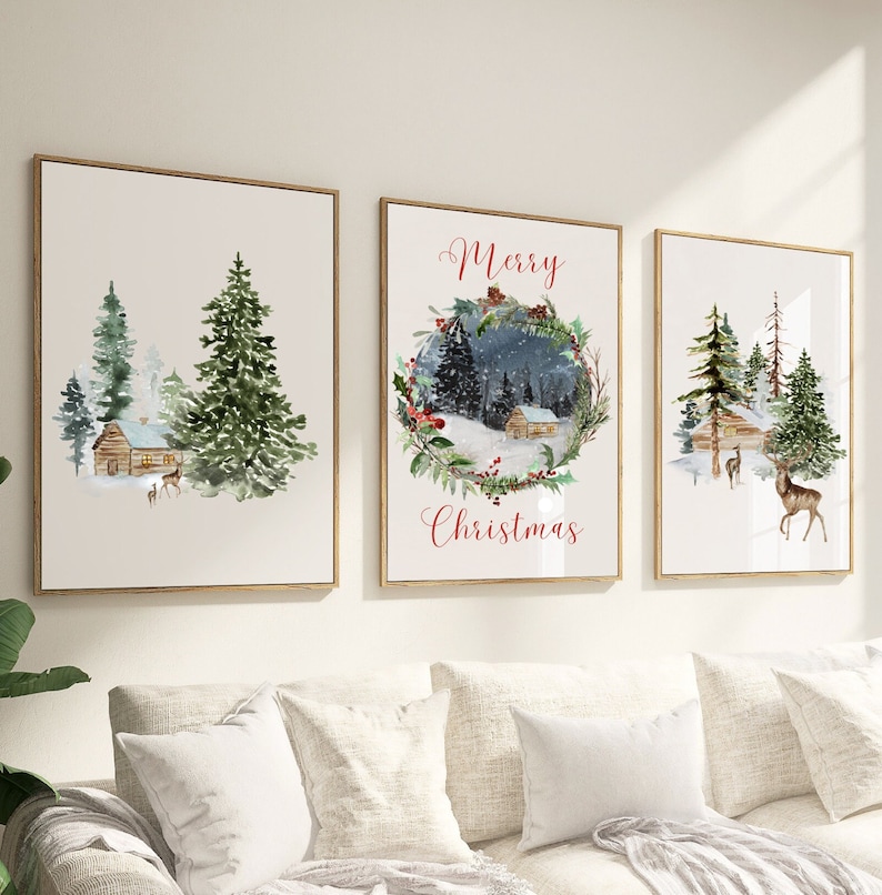 May include: Set of three framed watercolor prints featuring winter scenes. Each print has a gold frame and depicts a cabin, trees, and deer. One print has the text "Merry Christmas" in red script. The artwork evokes a cozy, festive atmosphere.