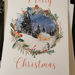 May include: A Christmas card with a watercolor painting of a snowy winter scene inside a wreath of greenery and berries. The words "Merry Christmas" are written in red script.