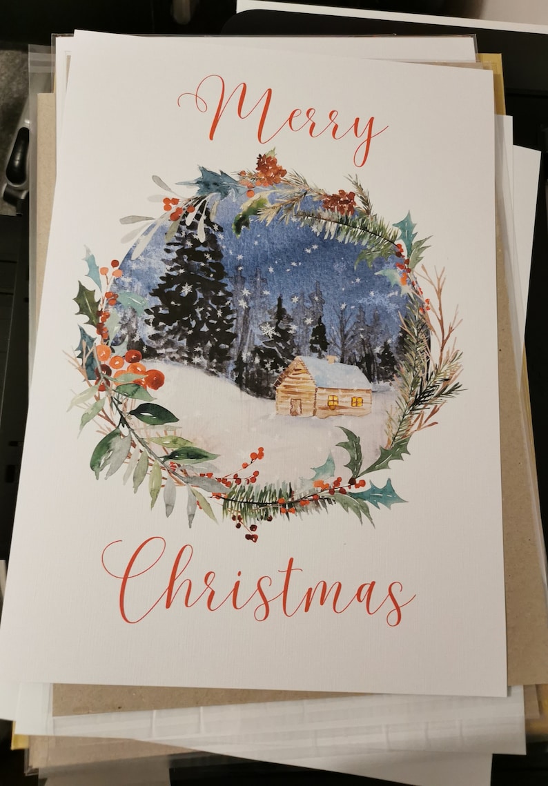 May include: A Christmas card with a watercolor painting of a snowy winter scene inside a wreath of greenery and berries. The words "Merry Christmas" are written in red script.