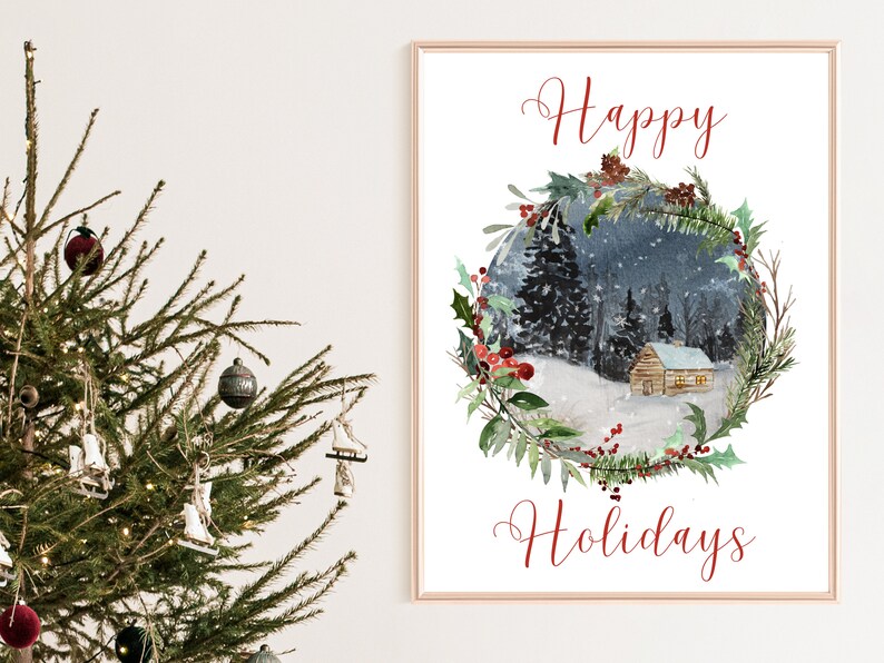 May include: A framed watercolor print with the words "Happy Holidays" in red script. The artwork depicts a snowy winter scene with a cabin nestled in a forest, surrounded by a wreath of greenery and red berries. A Christmas tree is in the foreground.
