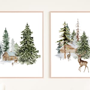 May include: Two framed watercolor prints depict winter scenes. One shows a cabin nestled among evergreen trees, with deer in the foreground. The other print features a similar cabin and trees, with deer. The artwork is in a soft, muted color palette.