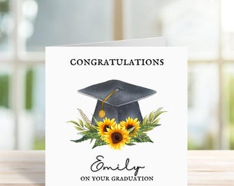 May include: A white graduation card with the word "CONGRATULATIONS" at the top. Below is a watercolor illustration of a black graduation cap, sunflowers, and greenery. The name "Emily" and the phrase "ON YOUR GRADUATION" are written in script.