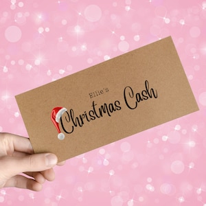 May include: A brown paper envelope with the text "Ellie's Christmas Cash" in black script, accompanied by a Santa hat illustration. The envelope is held against a pink background with white bokeh and starburst effects.