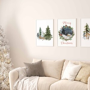 May include: Three framed watercolor prints with winter landscape scenes. The prints depict cabins, evergreen trees, and deer in a snowy setting. One print has the text "Merry Christmas" in red script. The prints are displayed above a beige sofa.