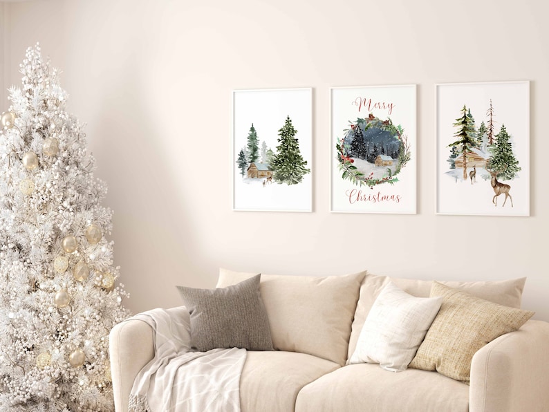 May include: Three framed watercolor prints with winter landscape scenes. The prints depict cabins, evergreen trees, and deer in a snowy setting. One print has the text "Merry Christmas" in red script. The prints are displayed above a beige sofa.