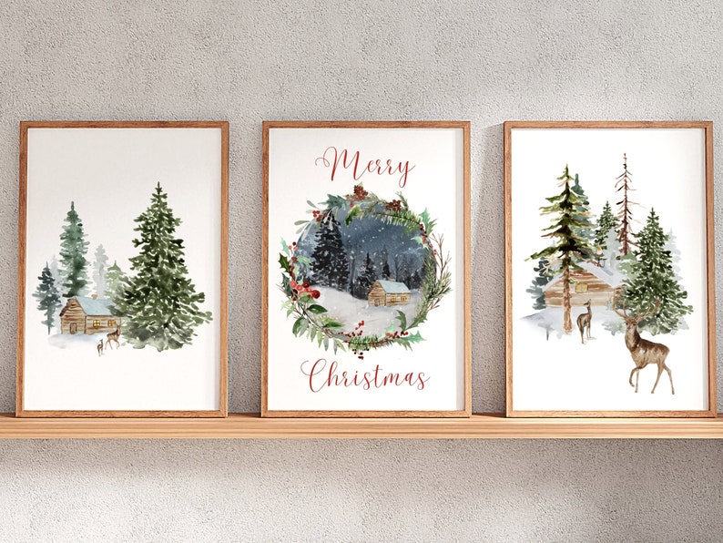 May include: Set of three framed watercolor prints featuring winter scenes. Each print depicts a cabin in a snowy forest with evergreen trees and deer. One print has a wreath with the words "Merry Christmas". The frames are a light brown color.