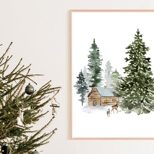 May include: Watercolor print of a winter scene with a cabin, evergreen trees, and deer. The artwork is framed in a gold-colored frame. A Christmas tree with ornaments is in the foreground.