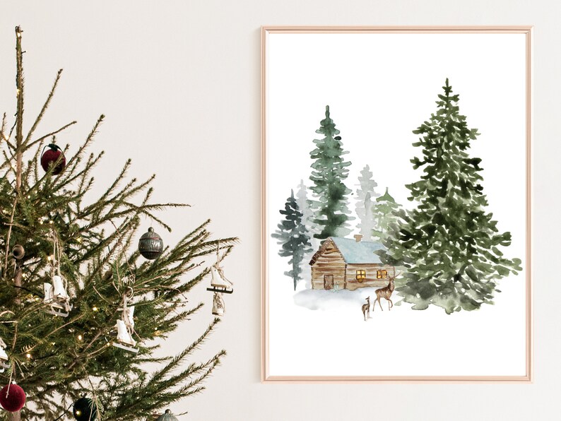 May include: Watercolor print of a winter scene with a cabin, evergreen trees, and deer. The artwork is framed in a gold-colored frame. A Christmas tree with ornaments is in the foreground.