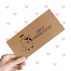 May include: A brown envelope with the text "Ellie's Holiday Funds" and a graphic of a suitcase with an airplane. The envelope is held against a white background with pink hearts.