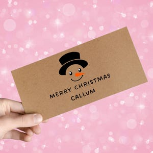 Peut inclure: Une carte de Noël marron avec un bonhomme de neige noir et orange et le texte "MERRY CHRISTMAS CALLUM". Le fond est un dégradé rose avec des étincelles blanches.