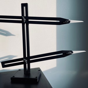 Op de afbeelding: Een zwart plastic gereedschap met twee witte messen gemonteerd op een standaard. De messen zijn verstelbaar en kunnen in verschillende posities worden geplaatst.