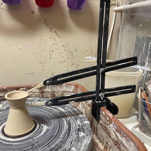 May include: A pottery wheel with a partially formed vase in the center. A black measuring tool is positioned over the vase. The wheel is surrounded by a brown basin, with a white bucket and other tools in the background.