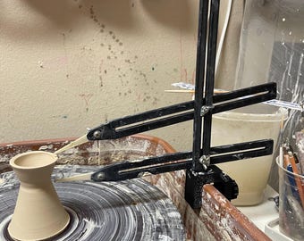 May include: A pottery wheel with a partially formed vase in the center. A black measuring tool is positioned over the vase. The wheel is surrounded by a brown basin, with a white bucket and other tools in the background.