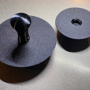 May include: Two black circular platforms with a textured surface. The smaller platform has a hole in the center. The larger platform has a black knob with a textured surface on top.
