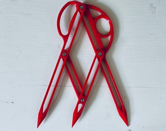 May include: A red plastic compass tool with two arms and a circular handle. The compass is designed for drawing circles and arcs.